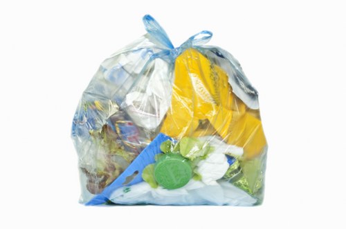A clear plastic rubbish bag filled with various household waste items, placed on a paved outdoor surface. The bag contains crumpled paper, plastic packaging, a yellow foam container, a green plastic bottle cap, and other mixed refuse materials, with the bag's semi-transparent plastic showing the contents inside. It is situated in an outdoor environment, possibly a driveway or street location in Berkhamsted, with no other objects or vehicles visible nearby. The bag appears to be loosely tied at the top, and the waste inside is pressed against the plastic, revealing an assortment of different textures and colours, including white, green, yellow, and blue. The lighting is natural, with even illumination highlighting the details of the bag and its contents, emphasizing the type of waste typically handled by a professional rubbish removal service such as Commercial Waste Berkhamsted. This scene exemplifies the type of waste collection and disposal work associated with local rubbish removal services in the area.