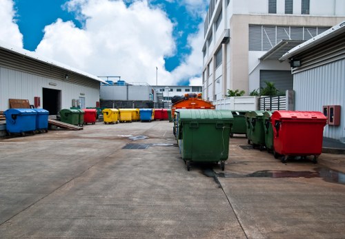 A large outdoor commercial waste collection area featuring multiple wheeled bins arranged in a semi-organised manner. The bins are made of plastic with textured surfaces and come in various colours including green, red, yellow, and blue, reflecting different waste types. They are positioned on a concrete surface within an industrial or business premises, with some bins leaning slightly or lined up along the edges. Behind the bins, there are white warehouse-style buildings with metal sidings, and the scene is set against a bright blue sky with scattered white clouds, suggesting daytime with natural sunlight illuminating the area. The environment appears clean but utilitarian, consistent with rubbish collection and waste management services in Berkhamsted or nearby local areas. The scene visually aligns with commercial waste removal services provided by Commercial Waste Berkhamsted, emphasizing the practical disposal of various waste materials at a business or industrial site in the region.