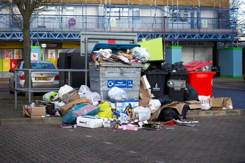 A white waste collection truck with an open rear loading compartment is parked on a narrow cobblestone street in an urban area. The vehicle appears to be engaged in rubbish removal, with the rear comp