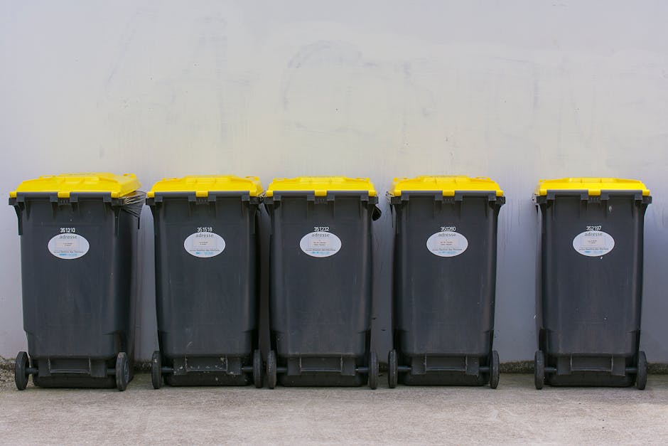 A person is disposing of crumpled brown paper bags and other waste items into a white rubbish bin with a grey lid, located on a wooden surface. The person's hand, visible and resting on the edge of th