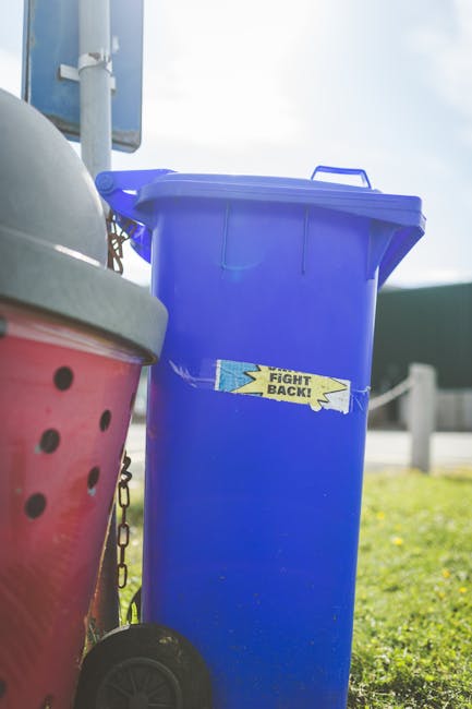 A tall, rectangular blue plastic rubbish bin with a closed lid, positioned outdoors on a grassy area. The bin features a small, rectangular sticker with the words 'FIGHT BACK!' in black text on a yell