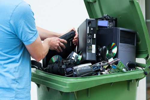 A person wearing a light blue t-shirt is standing beside a large green waste bin with its lid open, revealing a collection of discarded electronic waste items, including a damaged desktop computer tower, a black DSLR camera with a lens, several CDs and DVDs, and various cables and smaller electronic components. The setting appears to be outdoors on a paved surface, with natural daylight illuminating the scene. The electronic waste is haphazardly stacked within the bin, demonstrating typical electronic waste disposal practices. The image represents a professional approach to electrical and electronic waste collection, which involves proper rubbish removal services in Berkhamsted, Hertfordshire, offered by Commercial Waste Berkhamsted, targeting the management of domestic or office electronic waste through responsible disposal methods that support local waste regulations and environmental standards.
