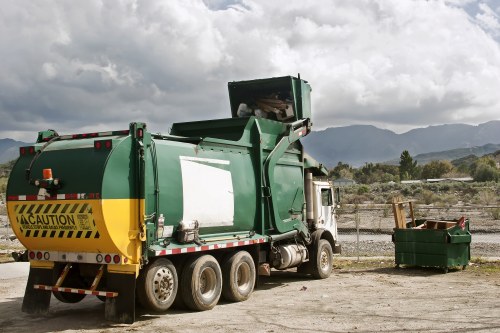 A large green waste collection truck with a yellow caution sign on the rear, parked on a gravel surface near a skip with some wooden pallets and other waste materials inside. The truck is positioned to the left, with the back end raised for dumping. In the background, there are mountains and a partly cloudy sky, indicating an outdoor setting typical of rubbish removal operations. The scene suggests a commercial waste collection service, possibly in or near a town such as Berkhamsted, with the vehicle ready for waste disposal or collection from a nearby premises. The environment appears functional and utilitarian, fitting for a professional rubbish removal service like Commercial Waste Berkhamsted, serving local businesses and communities in Hertfordshire.