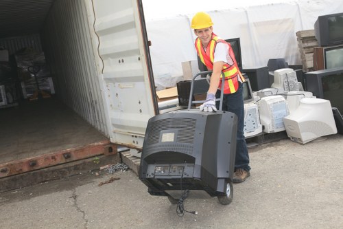 A person wearing a yellow safety helmet, high-visibility safety vest with pink and yellow stripes, and gloves is standing on an outdoor paved surface near the entrance of a storage container or warehouse. They are holding and guiding a portable CRT monitor, which is black and rectangular with visible buttons or ports on the front panel, and a cable hanging from the bottom. Behind them, there are stacks of various electronic waste items, including old computer towers and monitors, placed on the ground in front of a row of stacked pallets. The background shows a white covered structure and some bricks. This scene depicts a waste removal operation, likely involving the collection and disposal of electronic rubbish, and is relevant to commercial waste management services such as those offered by Commercial Waste Berkhamsted, serving clients in the Berkhamsted area within postcode HP4.